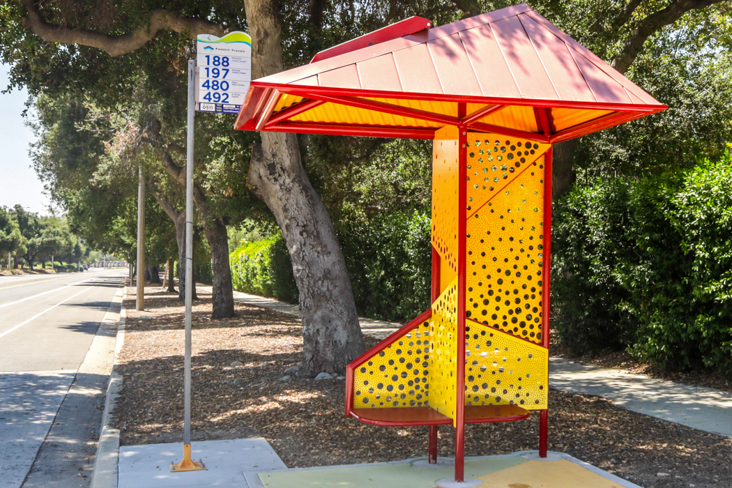 A red roof bus shelter with yellow hole design back paneling and two seats for riders is seen from the right side.