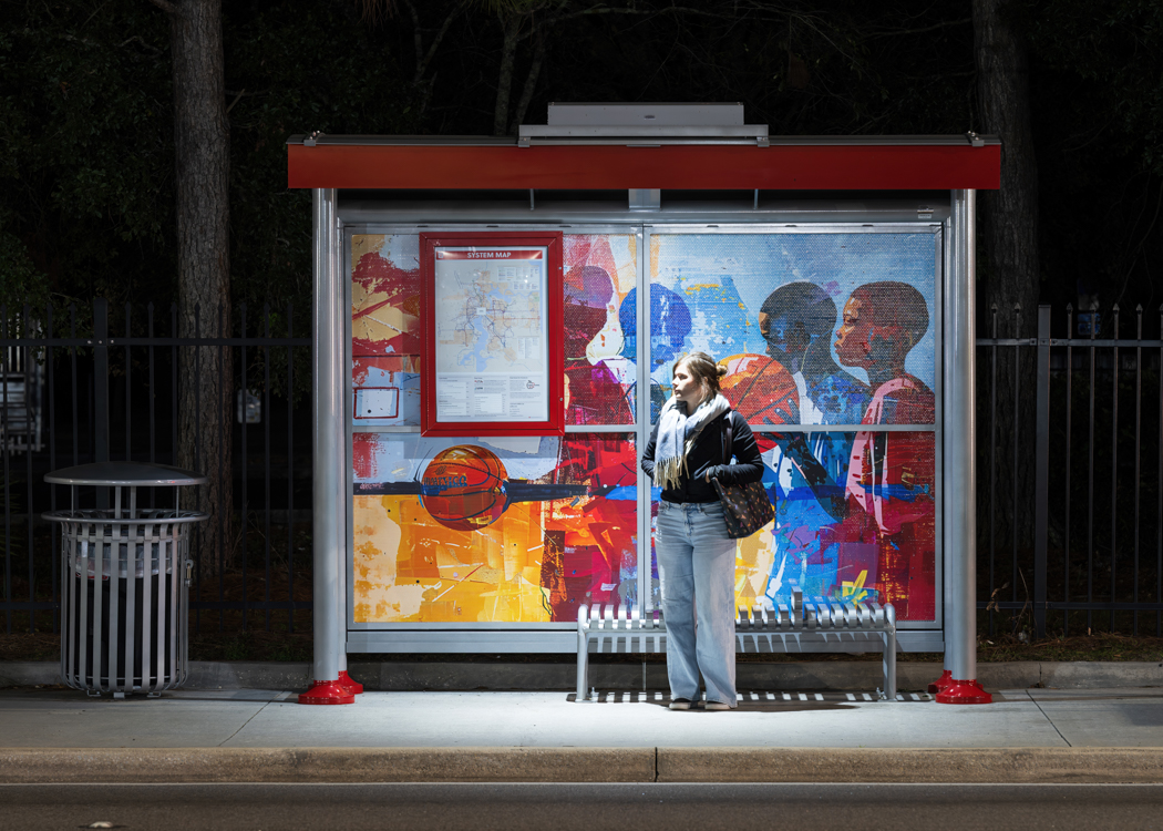 A bus shelter with a rider information display on the back panel and a rider waiting for the bus under the shelter at night with ample lighting.